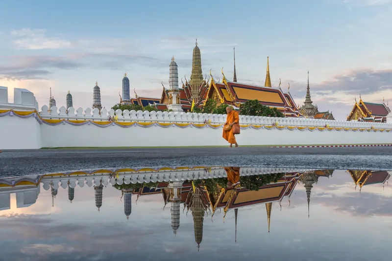 Gilded spires of the Temple of the Emerald Buddha rising behind the white Grand Palace wall, reflected in a wet courtyard, with a monk in an orange robe walking past.