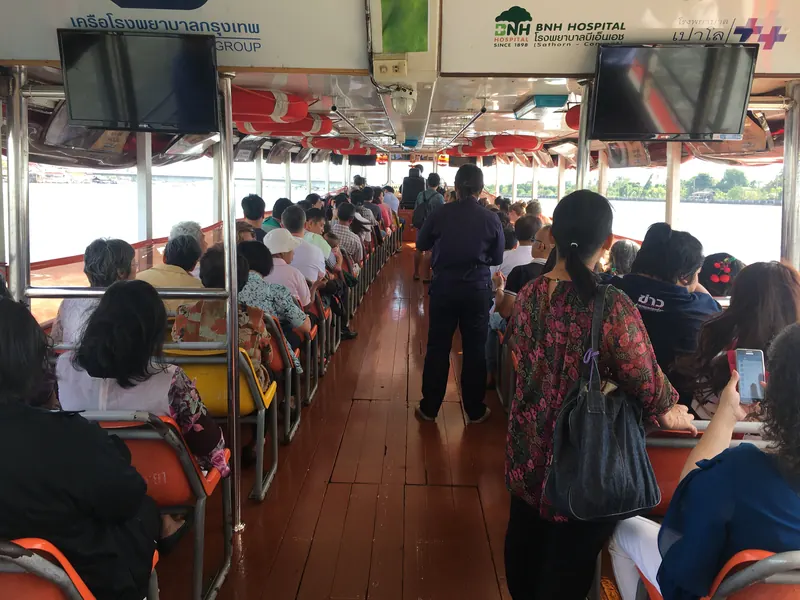 Passengers seated and standing inside a Chao Phraya Express Boat, with the river visible through open windows.