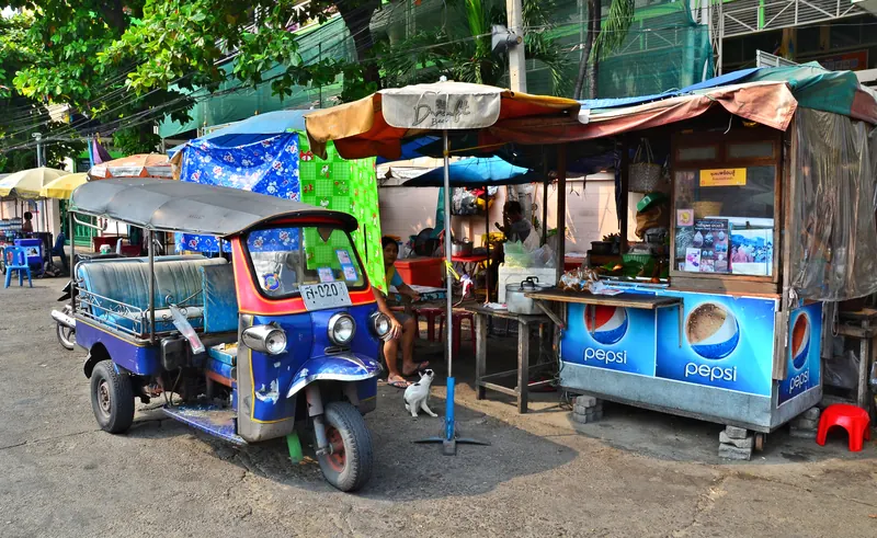 A blue and red tuk-tuk parked beside a street-food stall under canvas awnings in Bangkok.